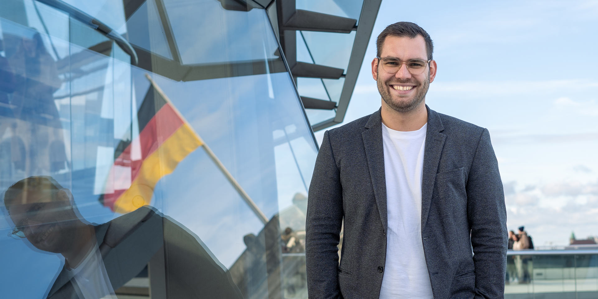 Daniel Baldy steht auf dem Dach des Bundestages, neben ihm spiegelt sich die Flagge der Bundesrepublik Deutschland im Fenster