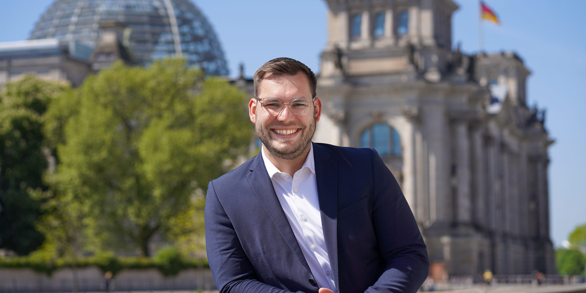 Daniel Baldy lächelt im Anzug, hinter ihm der Deutsche Bundestag